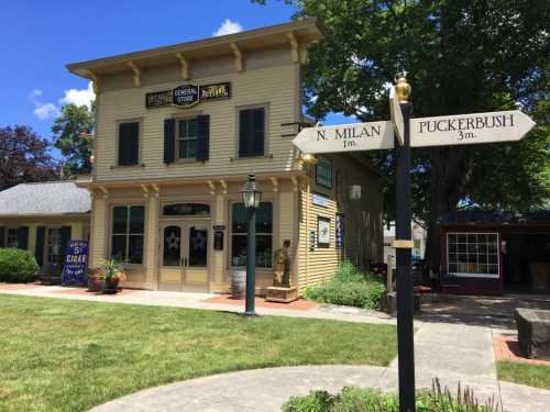 A charming yellow general store with a signpost indicating directions to North Milan and Puckerbush, set in a green landscape.