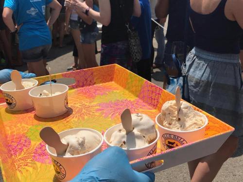 A hand holds a colorful tray with ice cream cups, while a crowd of people stands in the background on a sunny day.