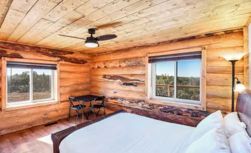 Cozy log cabin interior with a bed, small table, and large windows showcasing a scenic view.