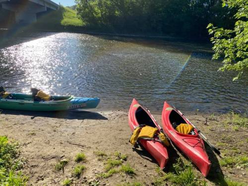 Two kayaks, one red and one blue, are parked on a sandy riverbank with sunlight reflecting on the water.