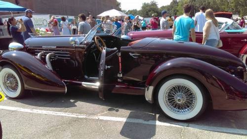 A classic maroon convertible car with whitewall tires, parked at a crowded outdoor event.