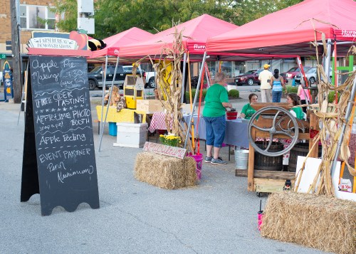 A farmers market scene with red tents, a chalkboard sign for apple pie and cider tasting, and people engaging in activities.
