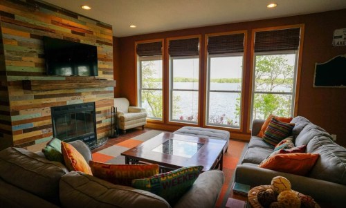 Cozy living room with large windows overlooking a lake, featuring a fireplace and comfortable seating.