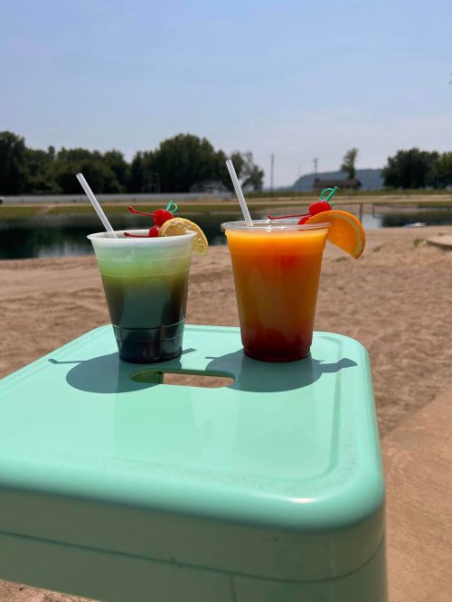 Two colorful drinks on a green table by a sandy area, with a lake in the background and a clear blue sky.