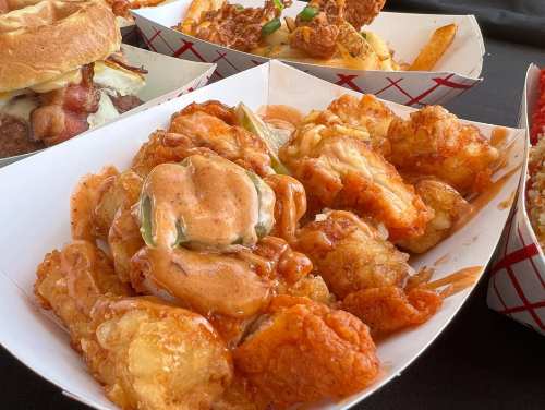 A close-up of crispy fried shrimp and tater tots drizzled with a spicy sauce, served in a paper tray.