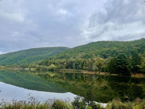 A serene lake reflecting green hills and cloudy skies, surrounded by lush trees and foliage.