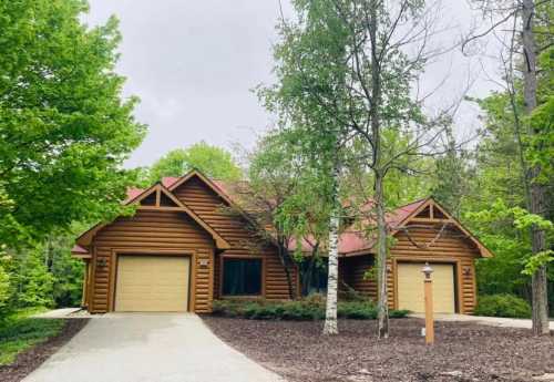 A log cabin-style house surrounded by lush green trees and a gravel driveway.