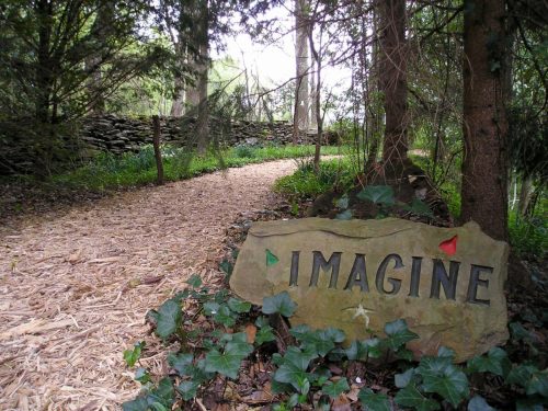 A stone sign reading "IMAGINE" beside a winding path through a lush, green forest.