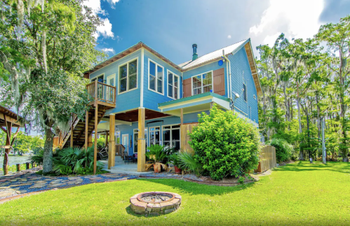 A blue two-story house surrounded by greenery, featuring a deck, fire pit, and views of a nearby waterway.