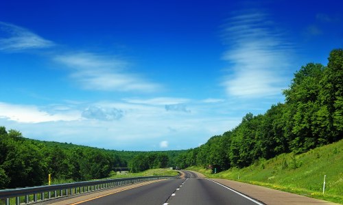A scenic highway stretches through lush green hills under a bright blue sky with scattered clouds.