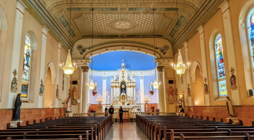 Interior of a church featuring ornate architecture, stained glass windows, and wooden pews, with two people standing at the altar.