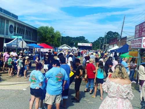 A bustling street fair with crowds, tents, and vendors under a clear blue sky. People are enjoying the festive atmosphere.