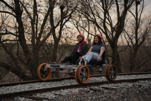 Two people sit on a rail cart on train tracks, surrounded by bare trees at dusk, both wearing helmets and smiling.
