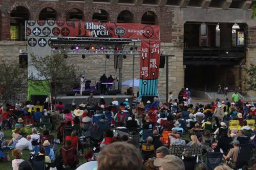 A large crowd enjoys a live music performance at a festival, with colorful banners and a stage in the background.