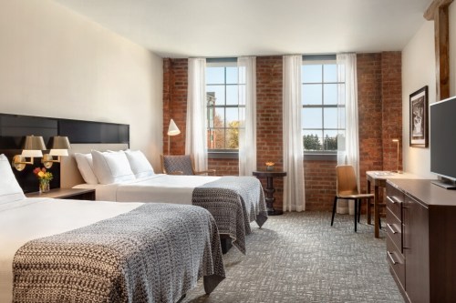 Bright hotel room with two beds, exposed brick walls, large windows, and modern furnishings.
