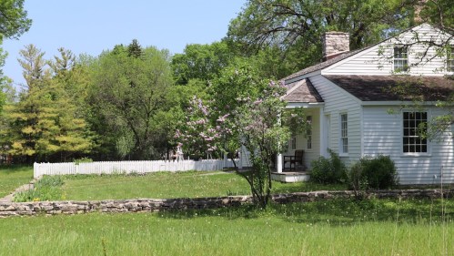 A white house with a porch surrounded by green grass, trees, and a flowering lilac bush, with a stone wall in the foreground.