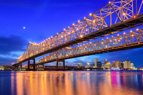 A brightly lit bridge spans a river at dusk, with city skyline reflections shimmering in the water below.