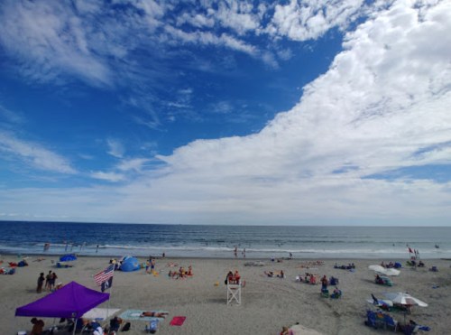 A beach scene with people relaxing on the sand, colorful umbrellas, and a blue sky with scattered clouds.
