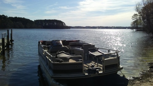 A pontoon boat is docked by a serene lake, with sunlight reflecting on the water and trees in the background.