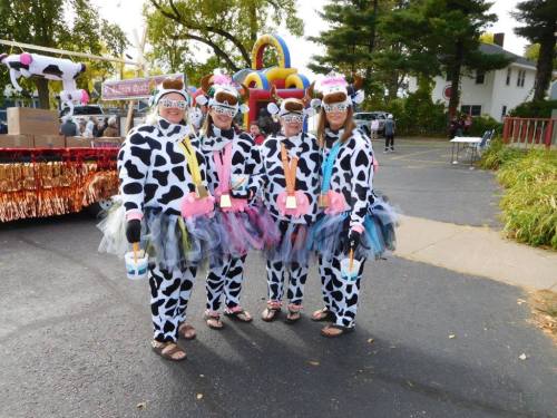 Four people in cow-print costumes with colorful tutus and accessories, posing at a festive outdoor event.