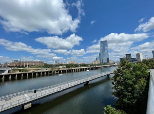 A scenic view of a river with a pedestrian bridge, city skyline, and fluffy clouds under a blue sky.