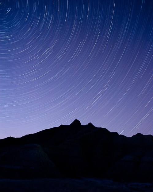 Star trails swirl above a dark mountain silhouette against a twilight sky.