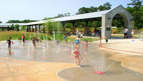 Children play in a splash pad, enjoying water fountains on a sunny day, with adults supervising nearby.