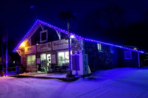 A stone building decorated with blue lights, surrounded by snow, at night. The sign reads "Witmer's Mercantile."