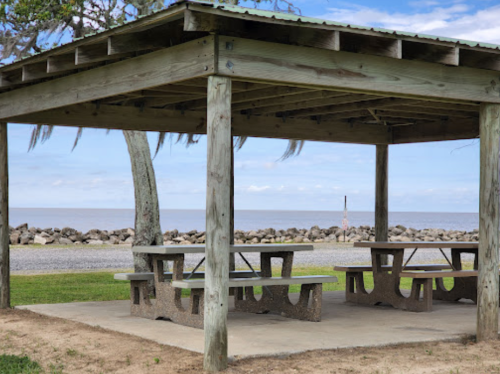 A wooden pavilion with picnic tables overlooking a calm body of water and a rocky shoreline under a blue sky.