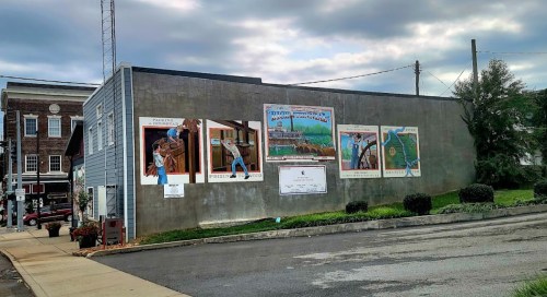 Mural on a building featuring colorful scenes and text, set against a cloudy sky in an urban area.