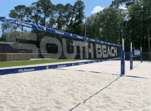 A beach volleyball court with a net displaying "SOUTH BEACH" in large letters, surrounded by trees.