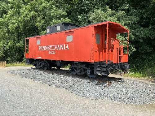 A red Pennsylvania caboose on a gravel track, surrounded by green trees.