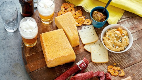 A wooden platter with various cheeses, pretzels, mustard, salami, and two glasses of beer.