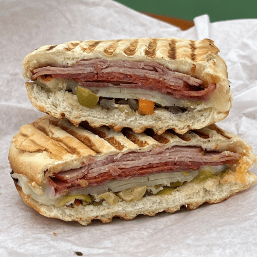 A sliced, grilled sandwich stacked with layers of meats, cheese, and pickles on toasted bread.