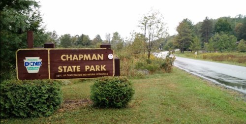 Sign for Chapman State Park, surrounded by greenery, with a road visible in the background.