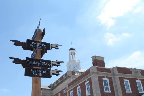 Signpost with directional arrows for events like Carnival and Bingo, with a historic building and clock tower in the background.