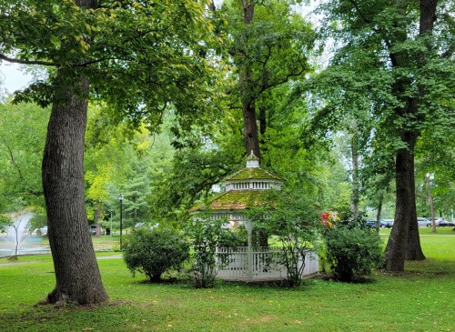A white gazebo surrounded by lush green trees and bushes in a peaceful park setting.