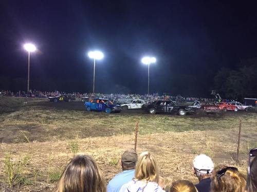 A night demolition derby with colorful cars colliding, surrounded by a large crowd under bright lights.