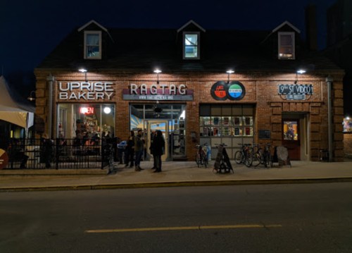 A brick building at night featuring Uprise Bakery and Ragtag, with people outside and bicycles parked nearby.