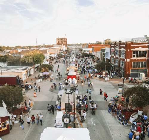A bustling street festival with crowds, food stalls, and shops lining the road under a clear sky.