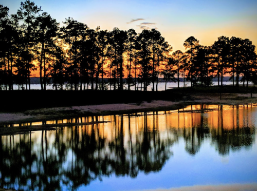 A serene lake at sunset, with trees silhouetted against a colorful sky and reflections on the water's surface.