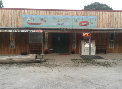 A rustic store with a wooden facade, offering kayaks, canoes, and snacks, featuring an "OPEN" sign and ice vending machine.