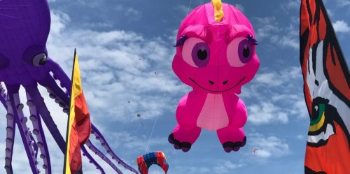 A large pink dinosaur kite with big eyes floats in a blue sky, surrounded by colorful flags and other kites.