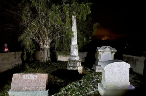 A dimly lit graveyard at night, featuring a tall obelisk and several gravestones surrounded by trees.