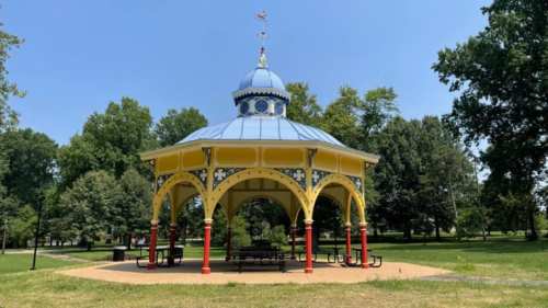 A colorful gazebo with a blue roof and yellow pillars, surrounded by trees in a sunny park.