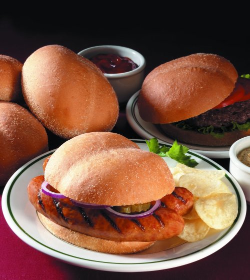A plate with a grilled sausage sandwich, potato chips, and soft rolls, with bowls of ketchup and mustard in the background.