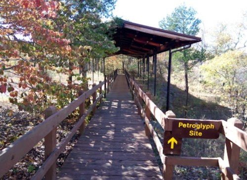 A wooden walkway leads to a covered area marked "Petroglyph Site," surrounded by trees and autumn foliage.
