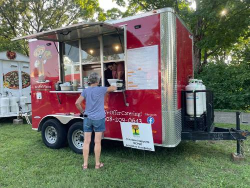 A person stands at a red food truck with a menu, surrounded by greenery. The truck offers catering services.