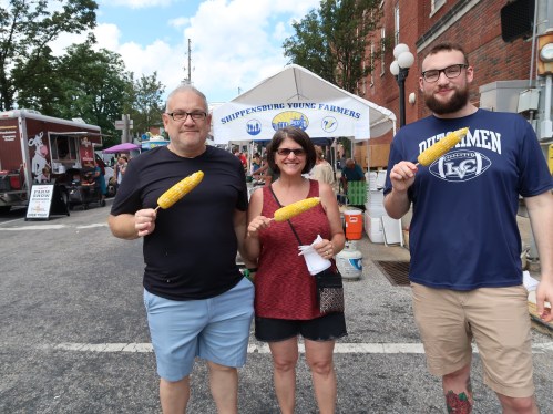 Three people hold corn on the cob in front of a food stand at an outdoor event. Bright day with trees in the background.