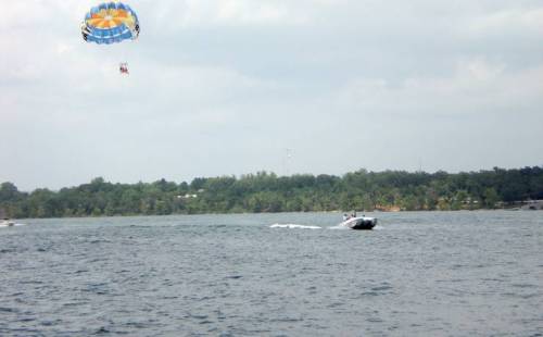 A boat speeds across a lake while a person paraglides above, with trees lining the shore in the background.